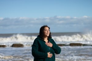 Amy with hand on heart and one on solar plexus
ocean and waves crash behind
peaceful expression 